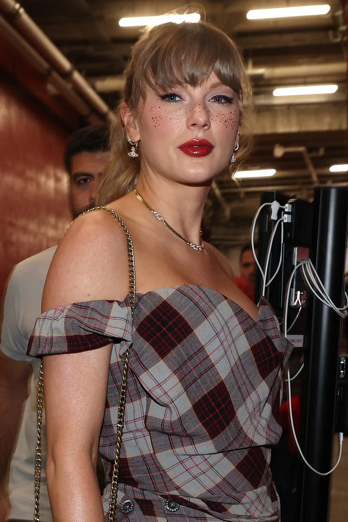 Taylor Swift egy focimeccsen kockás ruhában, piros rúzzsal és csillámos szeplőkkel.